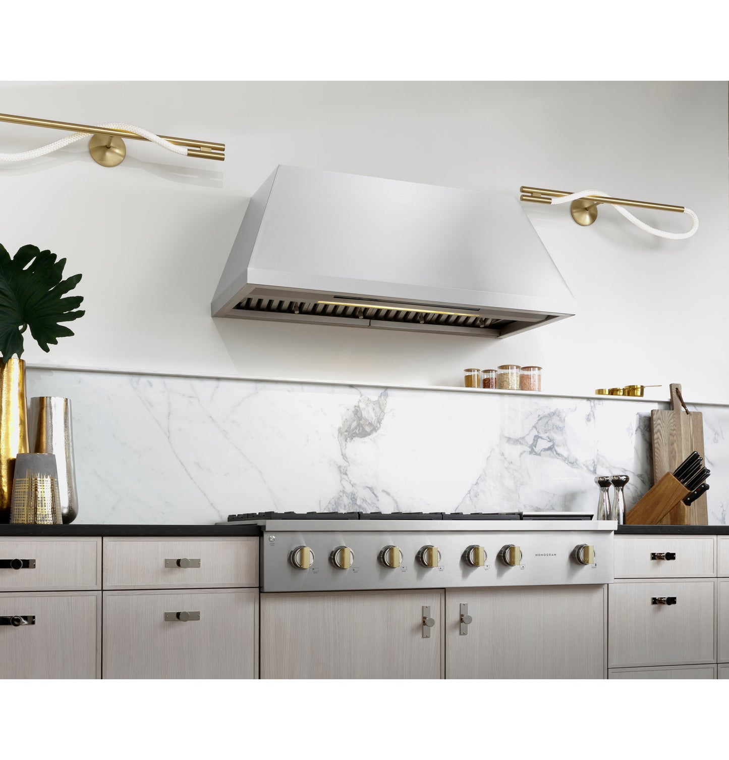Modern kitchen with stainless steel range hood above cooktop featuring six gold-tone control knobs and marble backsplash