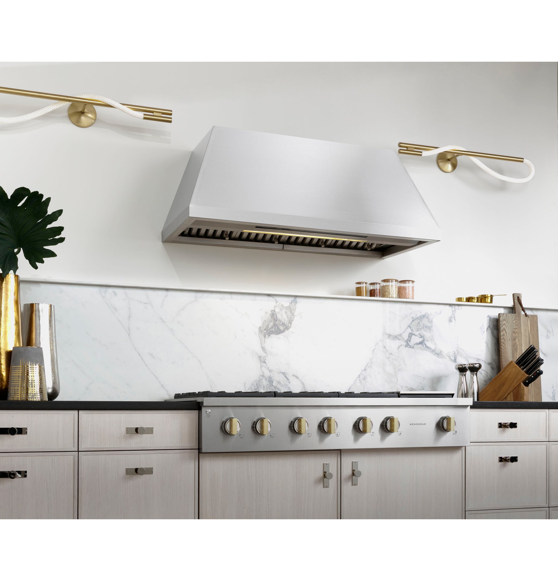 Modern kitchen with stainless steel range hood above cooktop featuring six gold-tone control knobs and marble backsplash