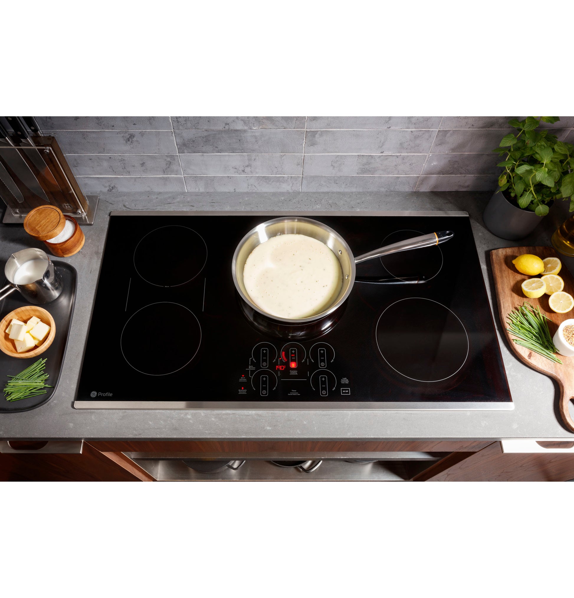 Modern black induction cooktop with touch controls heating a pot of sauce surrounded by fresh kitchen ingredients