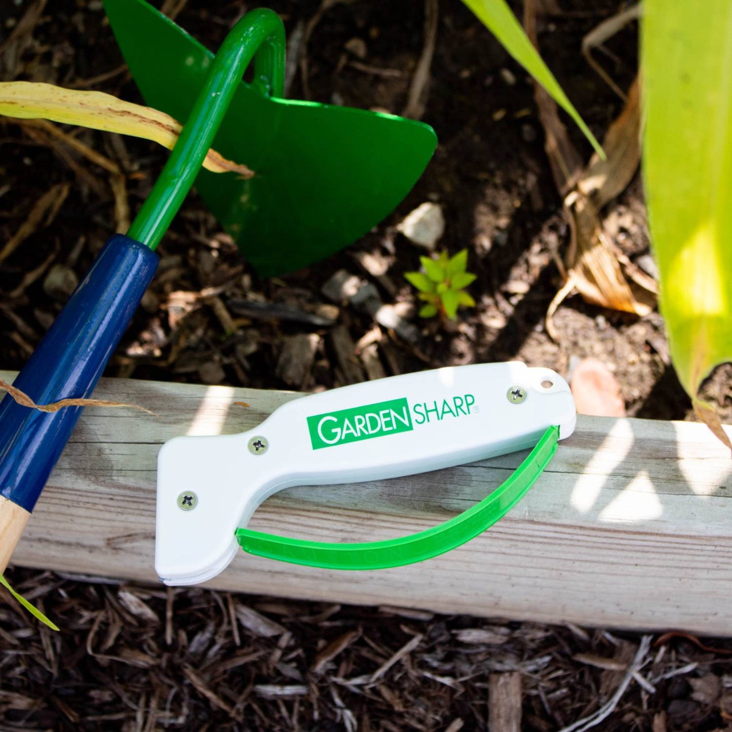 White and green handheld sharpener for gardening tools on a wooden garden bed with soil and a green hand hoe nearby