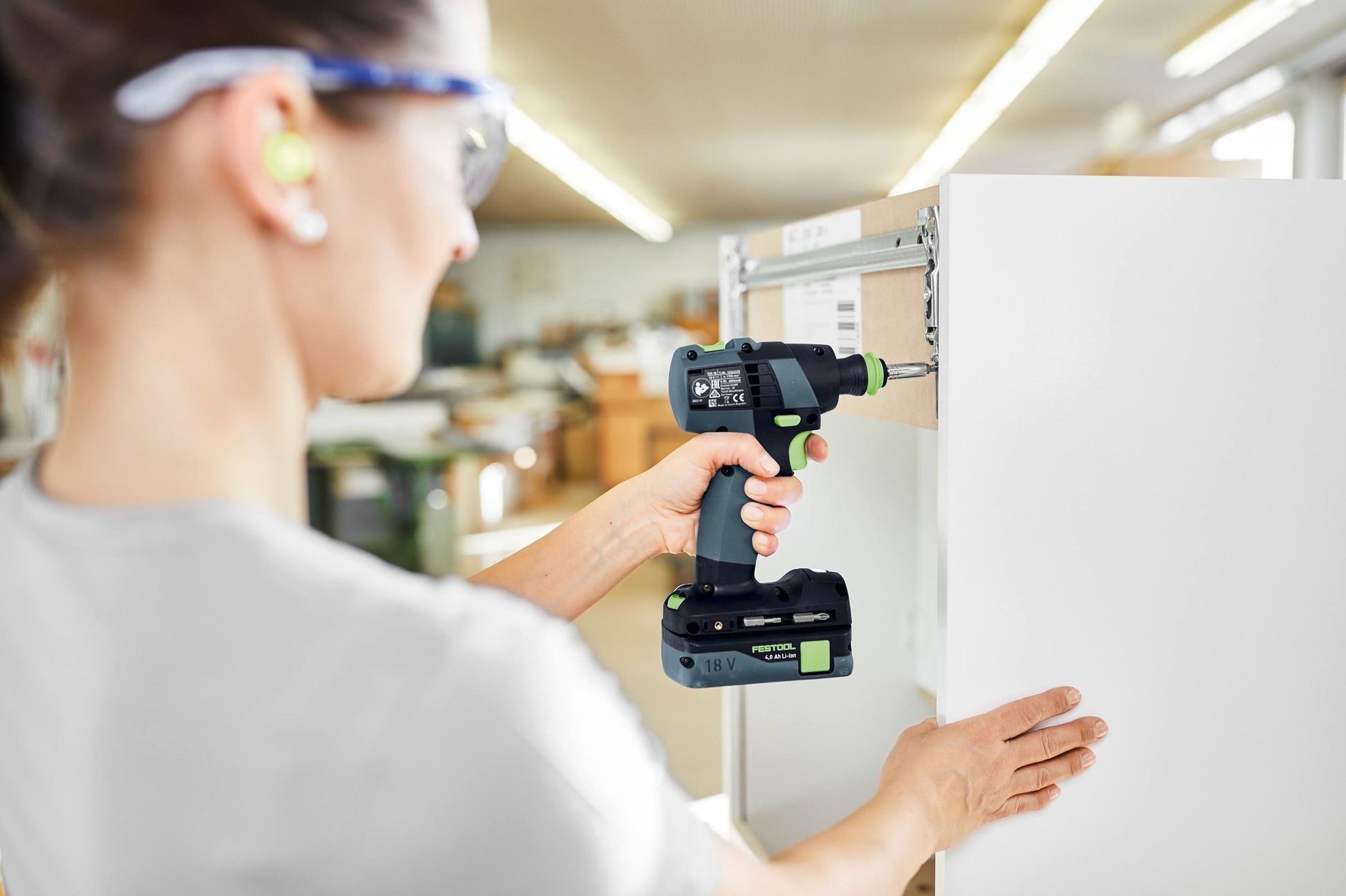 Woman wearing safety glasses and earplugs using compact cordless drill on white cabinet in workshop setting