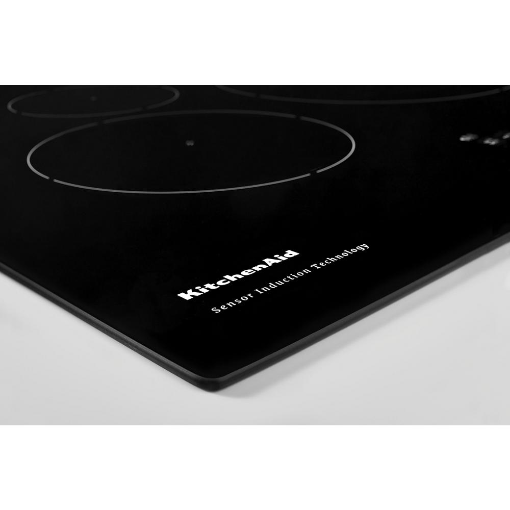 Close-up of black glass sensor induction cooktop corner with brand logo and smooth surface from KitchenAid collection