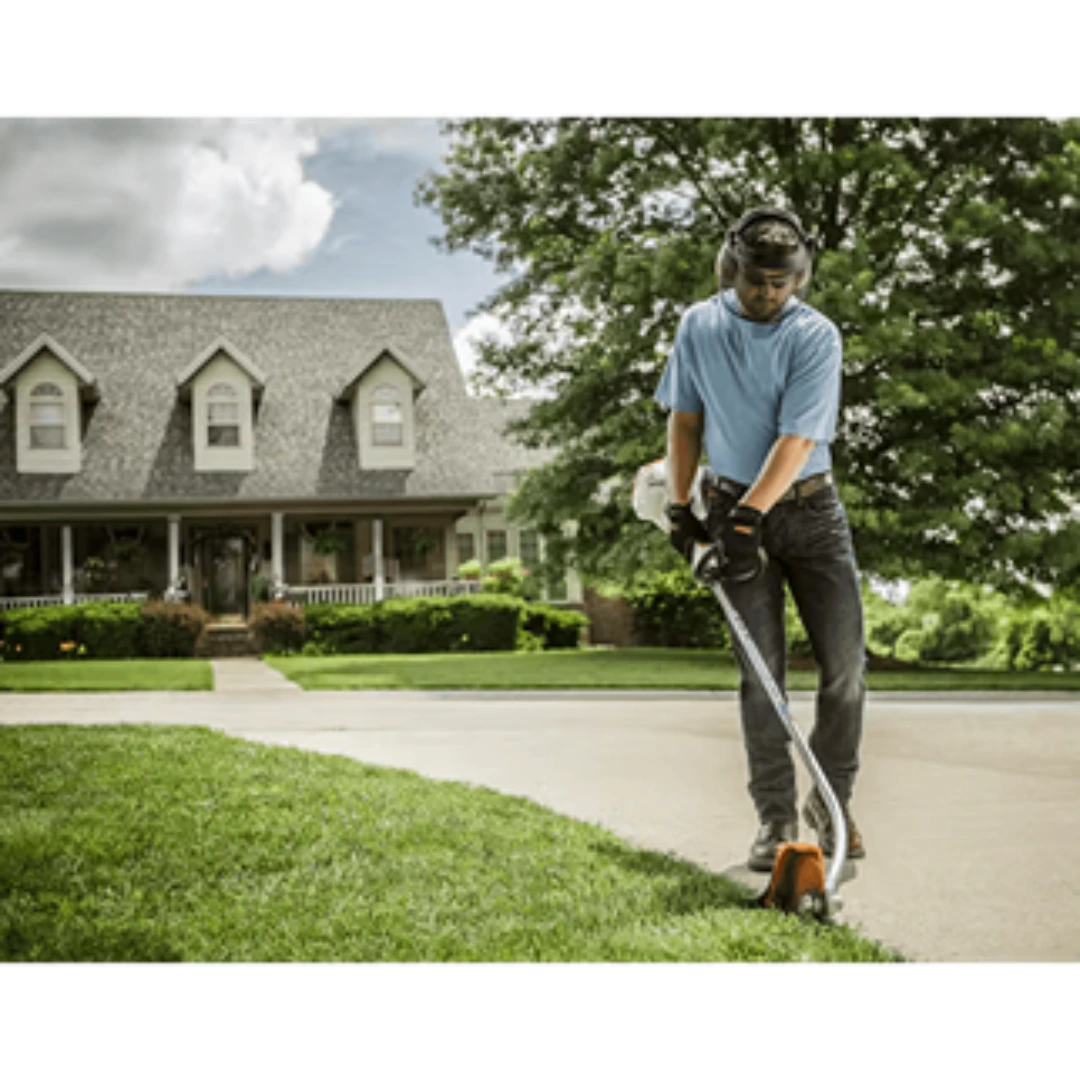 Man using a curved shaft edger to trim grass along a driveway in front of a suburban home for lawn care tasks