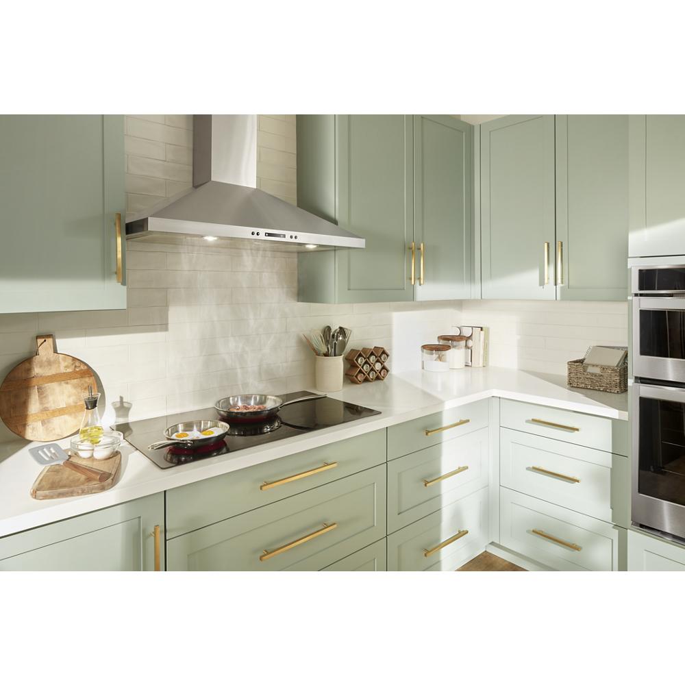 Modern kitchen corner with sage green cabinets, gold handles, stainless steel chimney range hood, and built-in double oven appliance