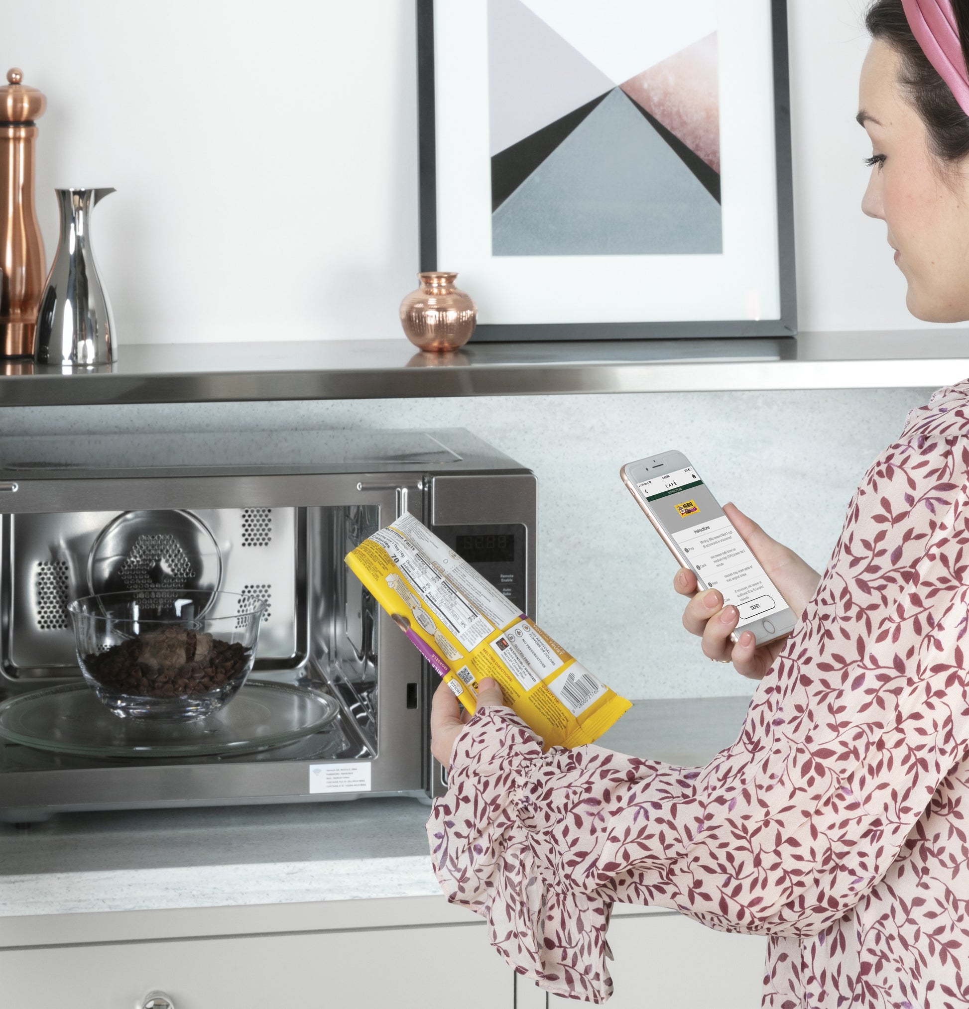 Woman using smartphone to control smart countertop microwave in modern kitchen with decorative items and artwork