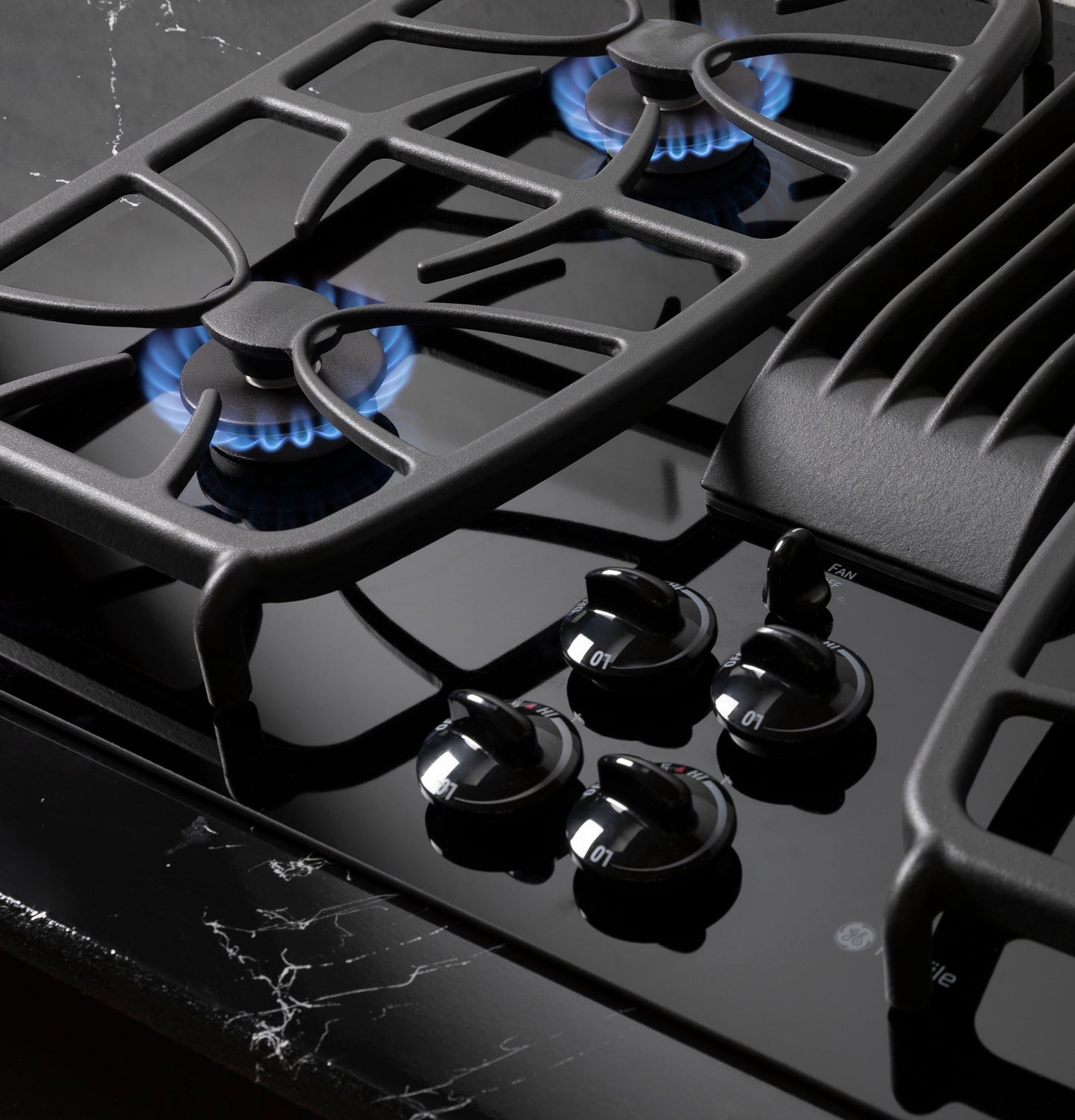 Close-up of black gas cooktop burners with blue flames and control knobs in a modern kitchen appliance setting