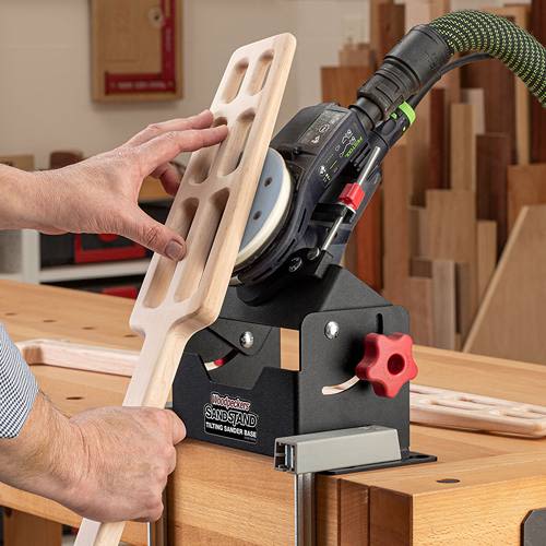 Black metal tilting sander base with red knob holding palm sander sanding carved wooden paddle tool for woodwork
