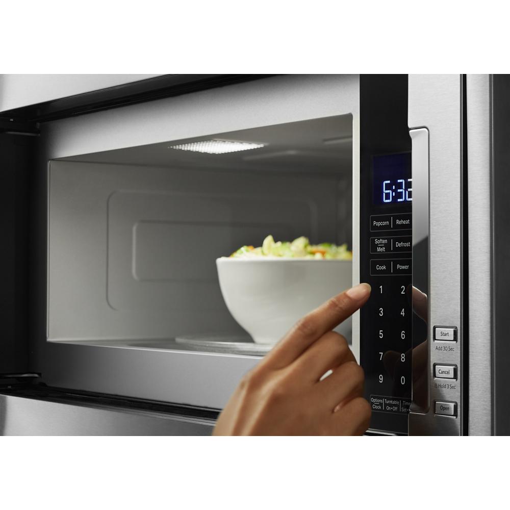 Hand pressing digital control panel on stainless steel built-in microwave with bowl of food inside kitchen appliances