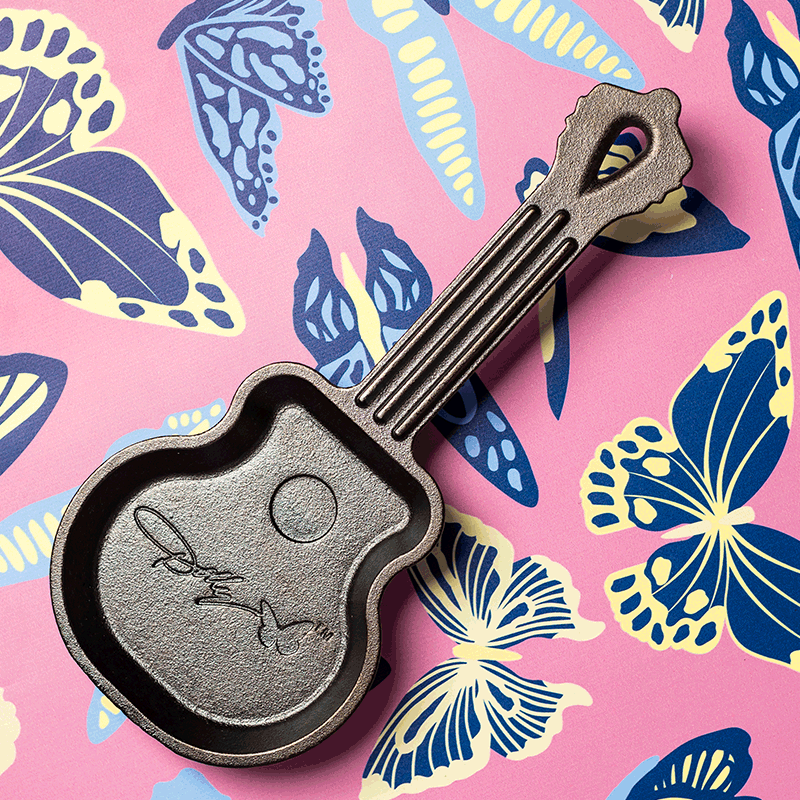 Cast iron guitar-shaped mini skillet with engraved signature detail resting on a pink and blue butterfly pattern background
