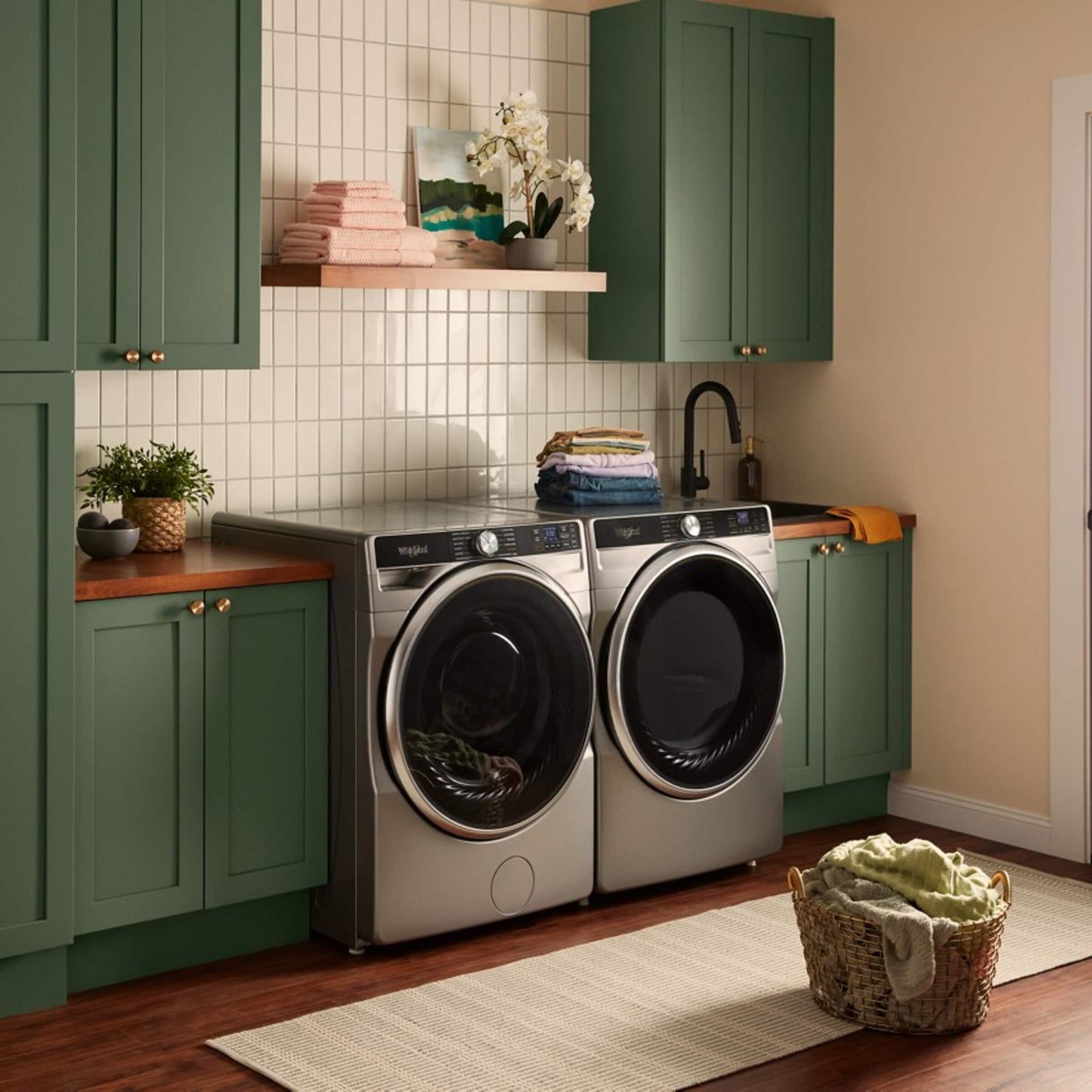 Laundry room with silver front load washer and dryer set surrounded by green cabinets and countertop space