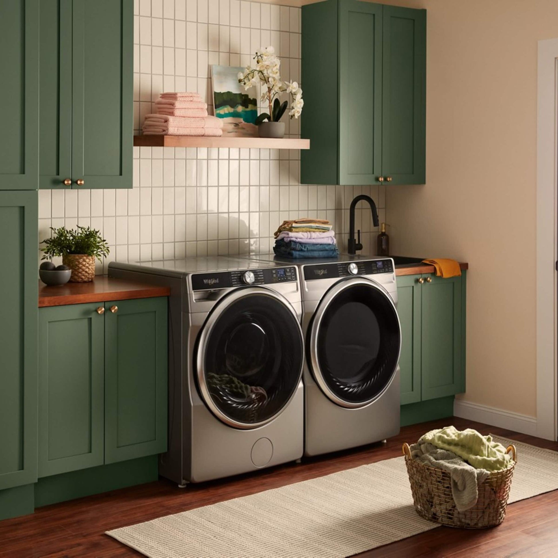 Laundry room with silver front load washer and dryer set surrounded by green cabinets and countertop space