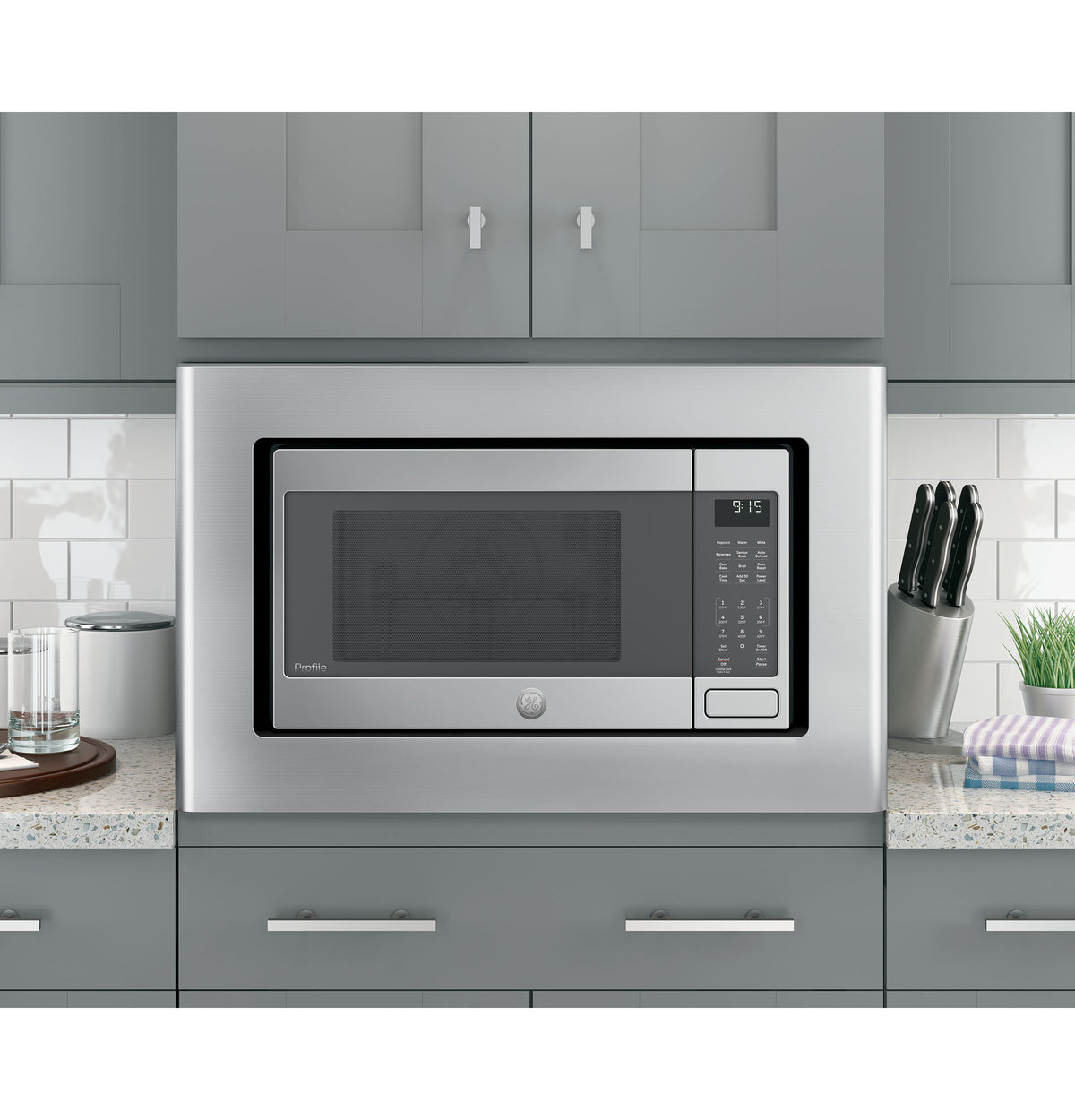 Stainless steel microwave trim kit installed in modern kitchen with gray cabinets and countertop accessories