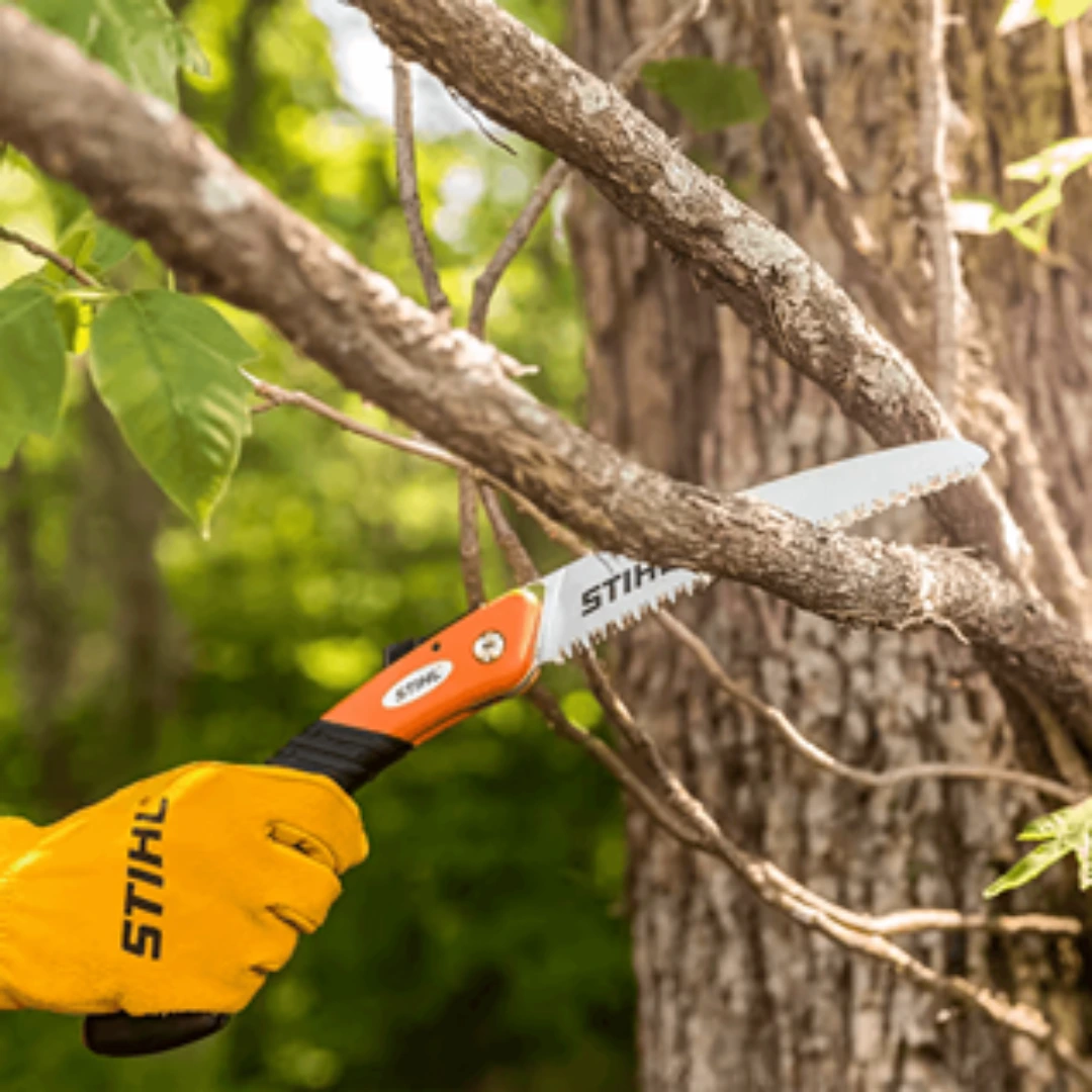 Compact folding saw with curved blade cutting tree branches, shown with STIHL gloves for outdoor power tools and lawn care