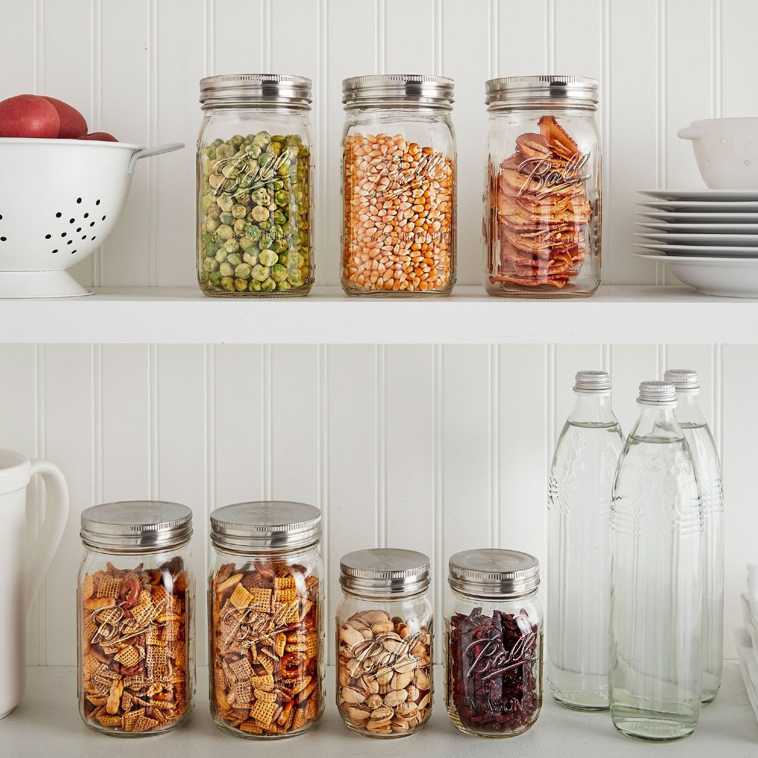 Seven clear glass jars with stainless steel lids containing various dried foods, arranged on white shelves in a kitchenware setting