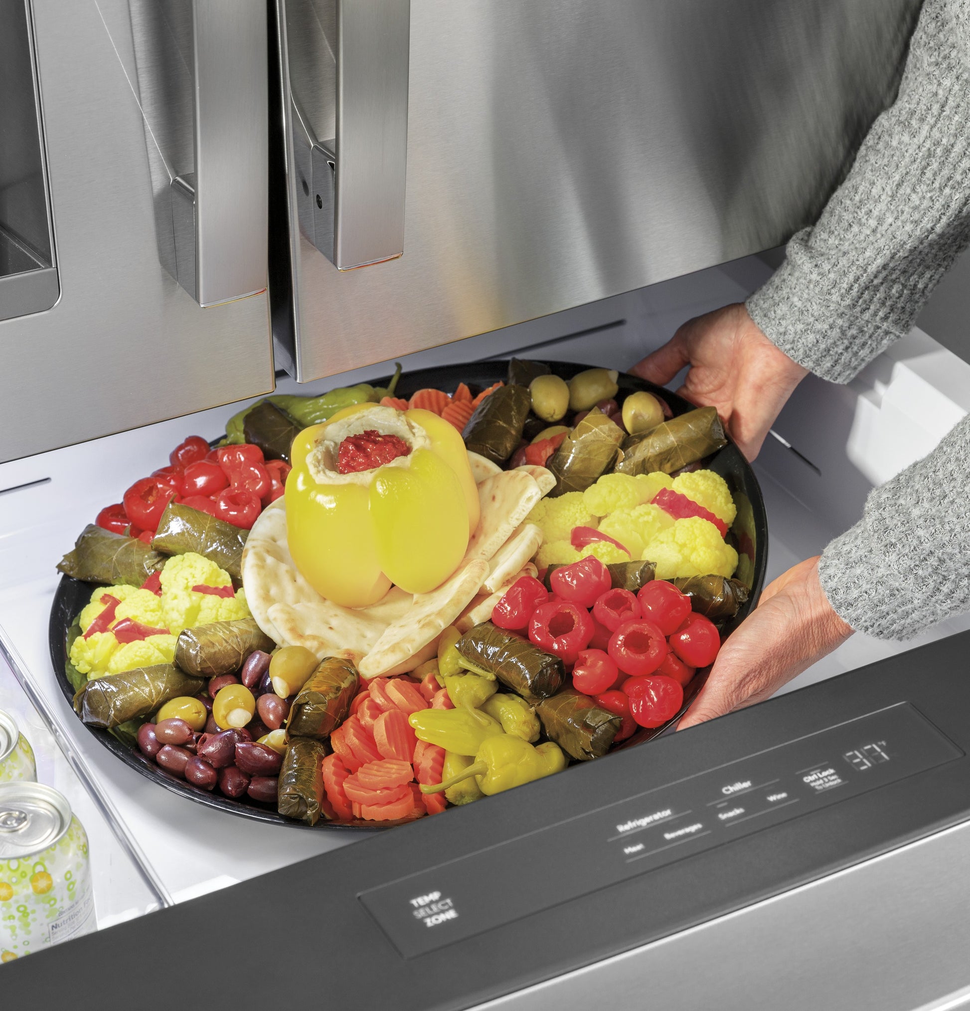 Person placing large platter of assorted appetizers including stuffed pepper and vine leaves inside bottom freezer of stainless steel French-door refrigerator