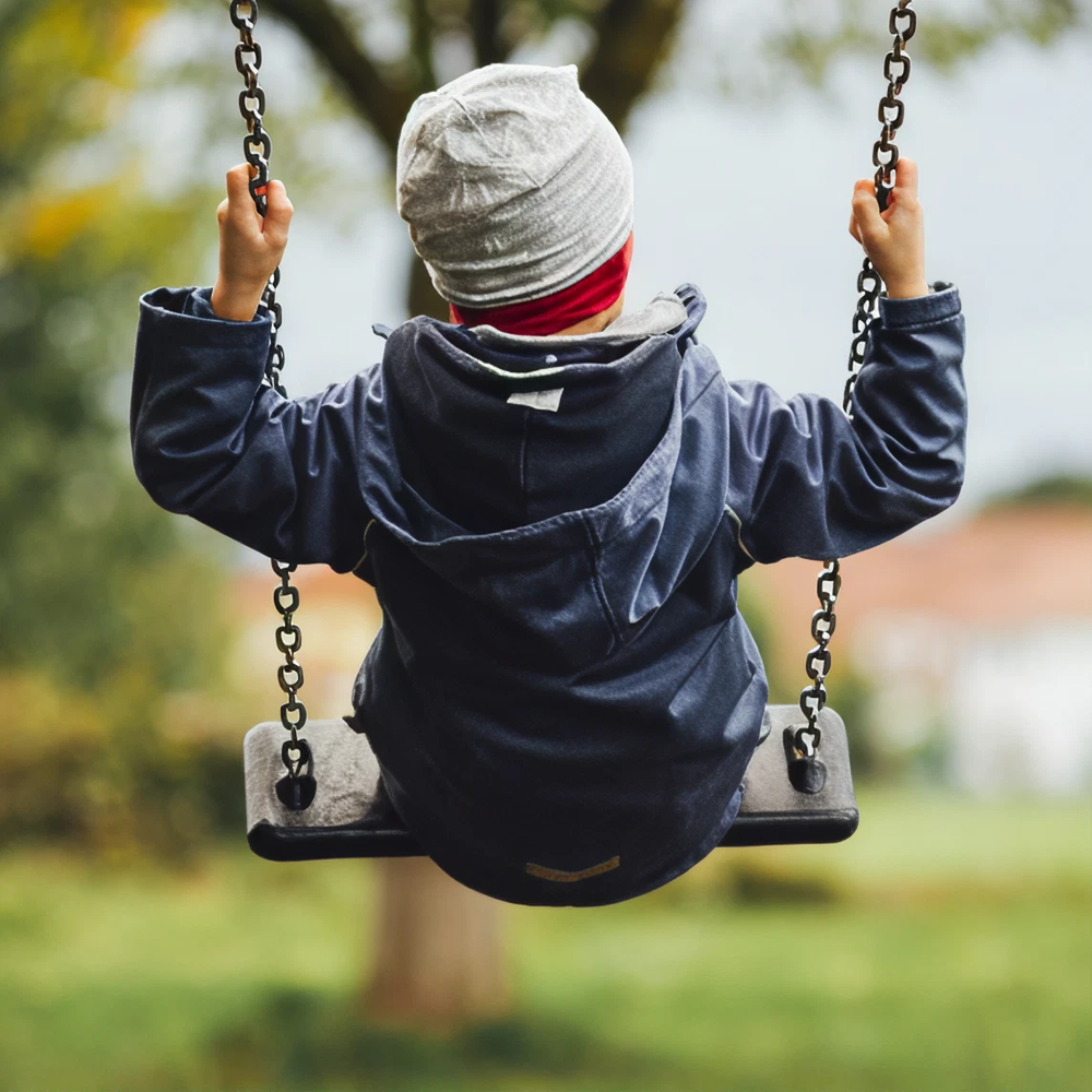 Child wearing a gray hat and navy jacket sitting on a swing outdoors, holding onto the chains with a blurred green background.