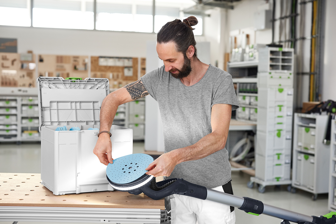 Man loading blue abrasive disc into long-reach sander with white storage box in workshop setting
