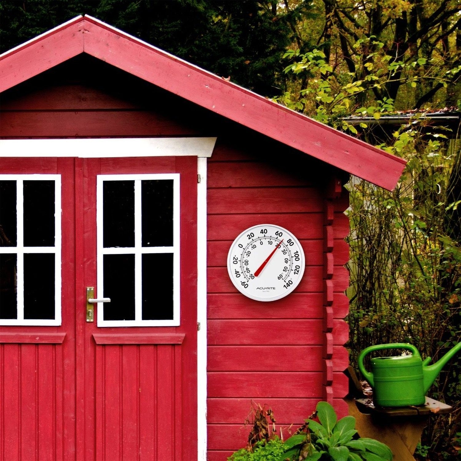 Outdoor round thermometer with white dial and red needle displaying temperature in Fahrenheit and Celsius on red wooden shed wall