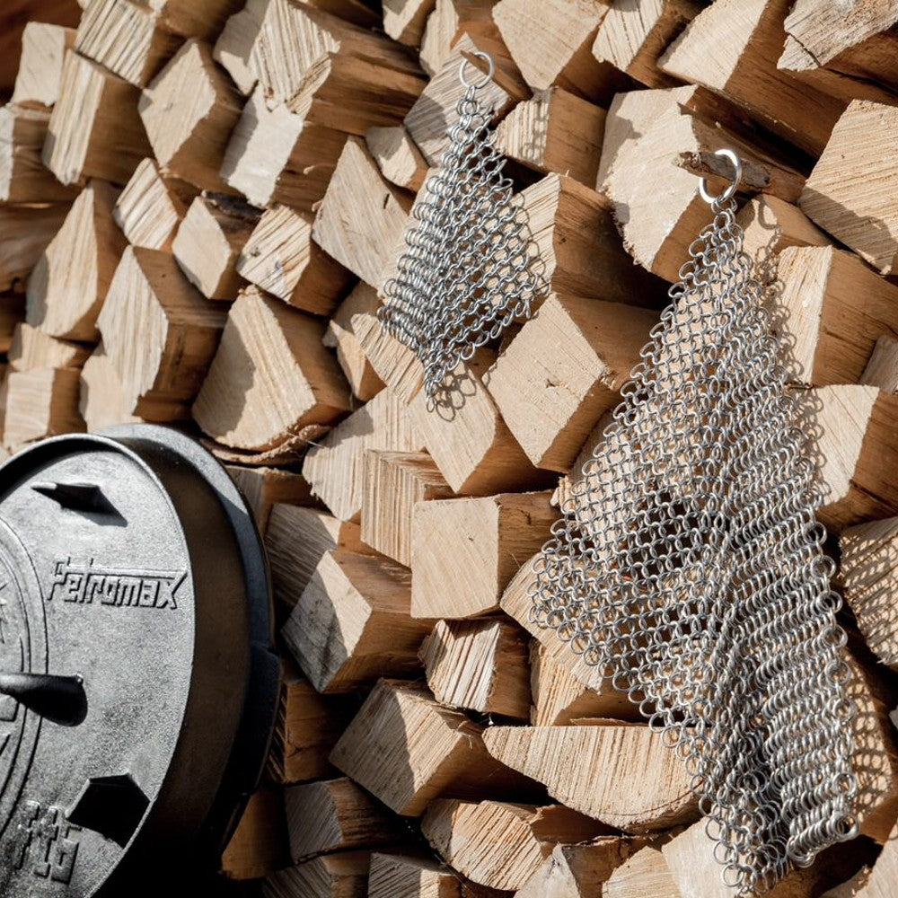 Two stainless steel chainmail scrubbers hanging on stacked firewood near cast iron lid in kitchenware collection under $50