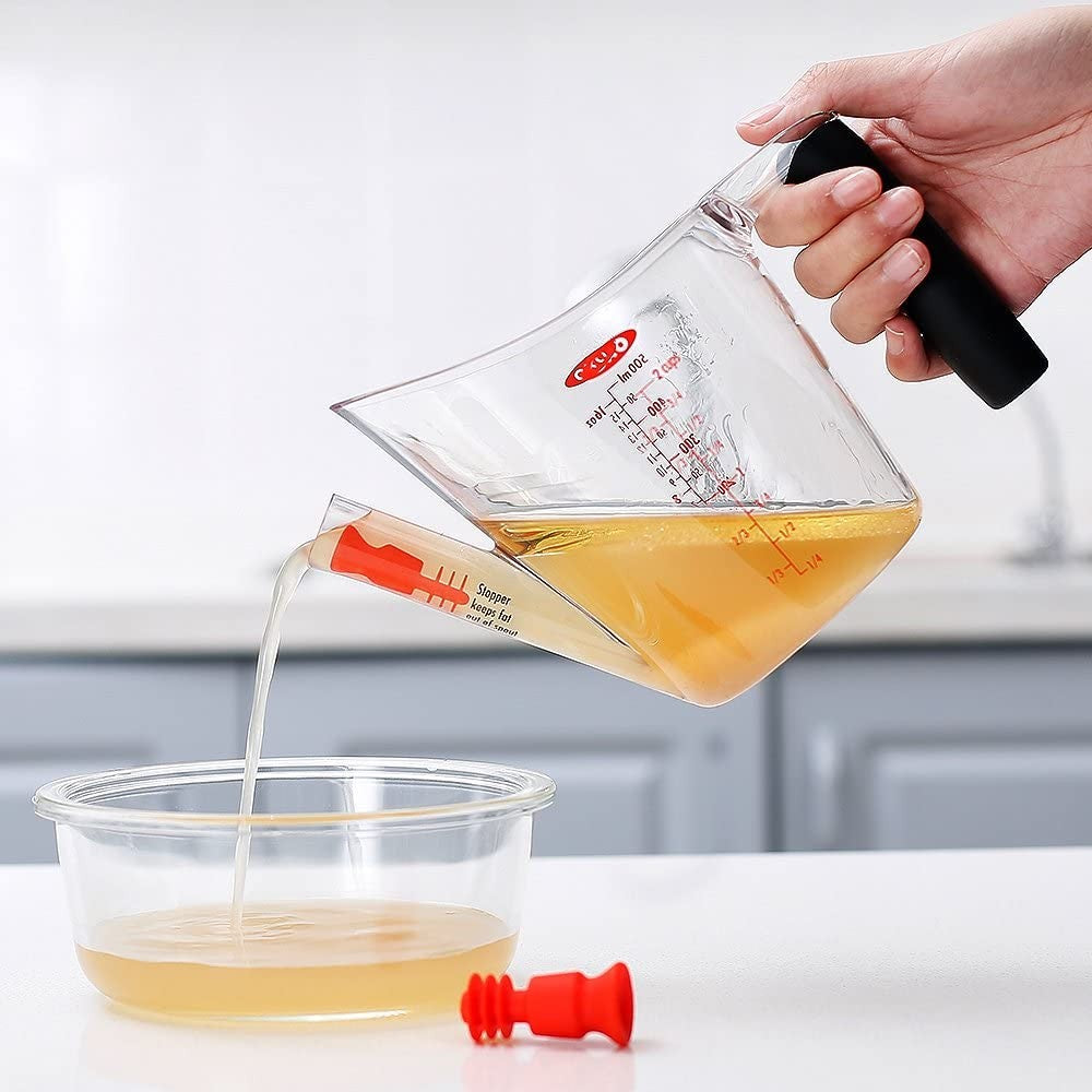 Clear OXO fat separator with black handle pouring broth through red stopper into clear bowl in kitchen gadgets collection