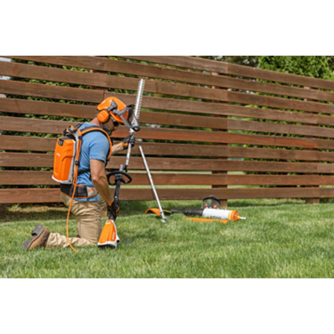 Worker wearing protective gear operating orange battery-powered KombiMotor tool on grass near wooden fence in outdoor power equipment setting