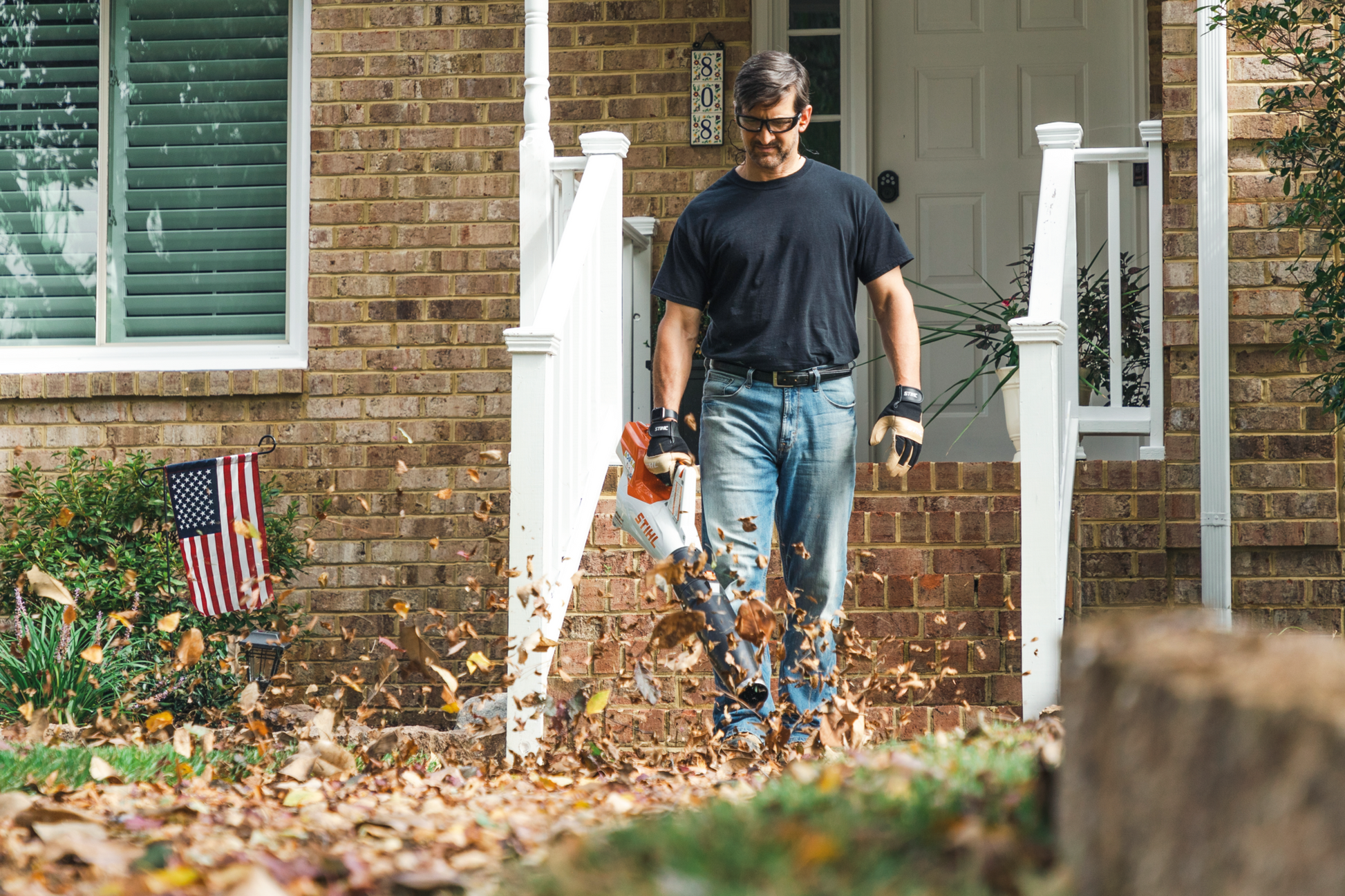 Man using a STIHL battery handheld blower to clear fallen leaves from a residential front yard during home improvement tasks