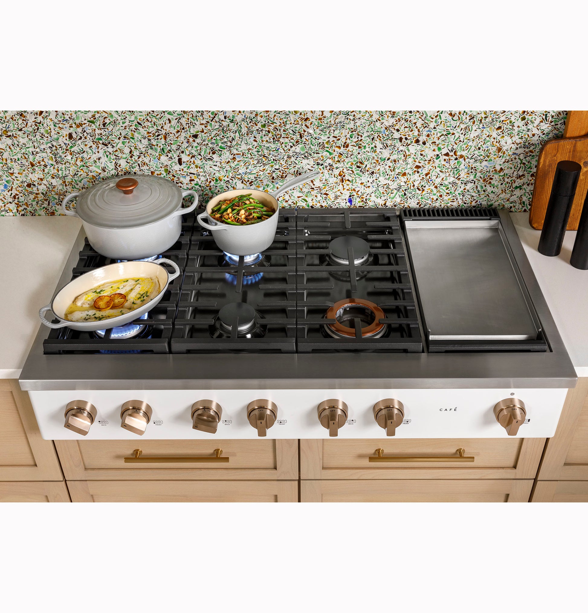 White gas rangetop with six burners and integrated griddle cooking dishes on light wood cabinetry in kitchenware collection