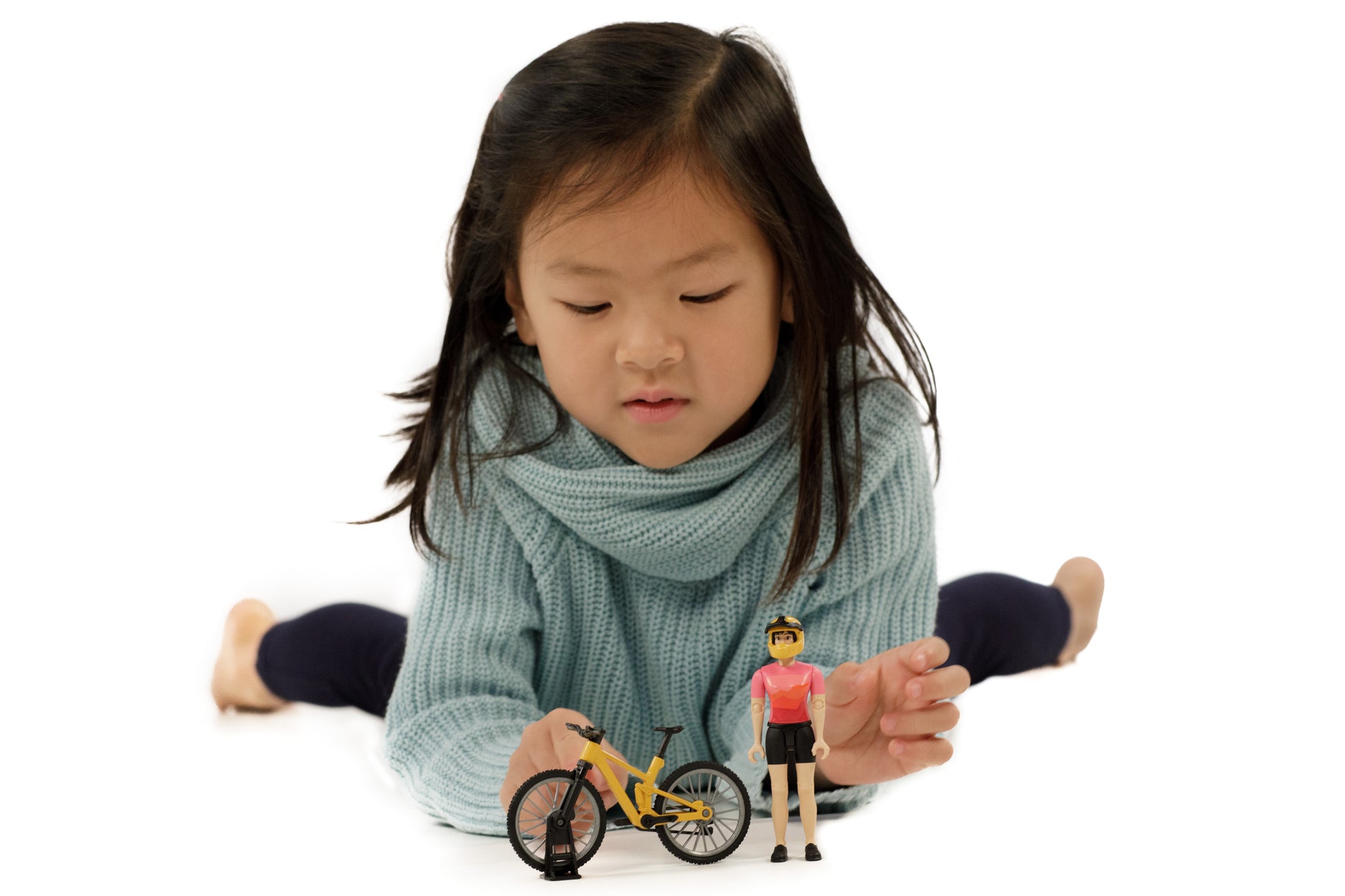 Young girl lying on floor playing with yellow bicycle toy and action figure from Bruder Toys collection under $50