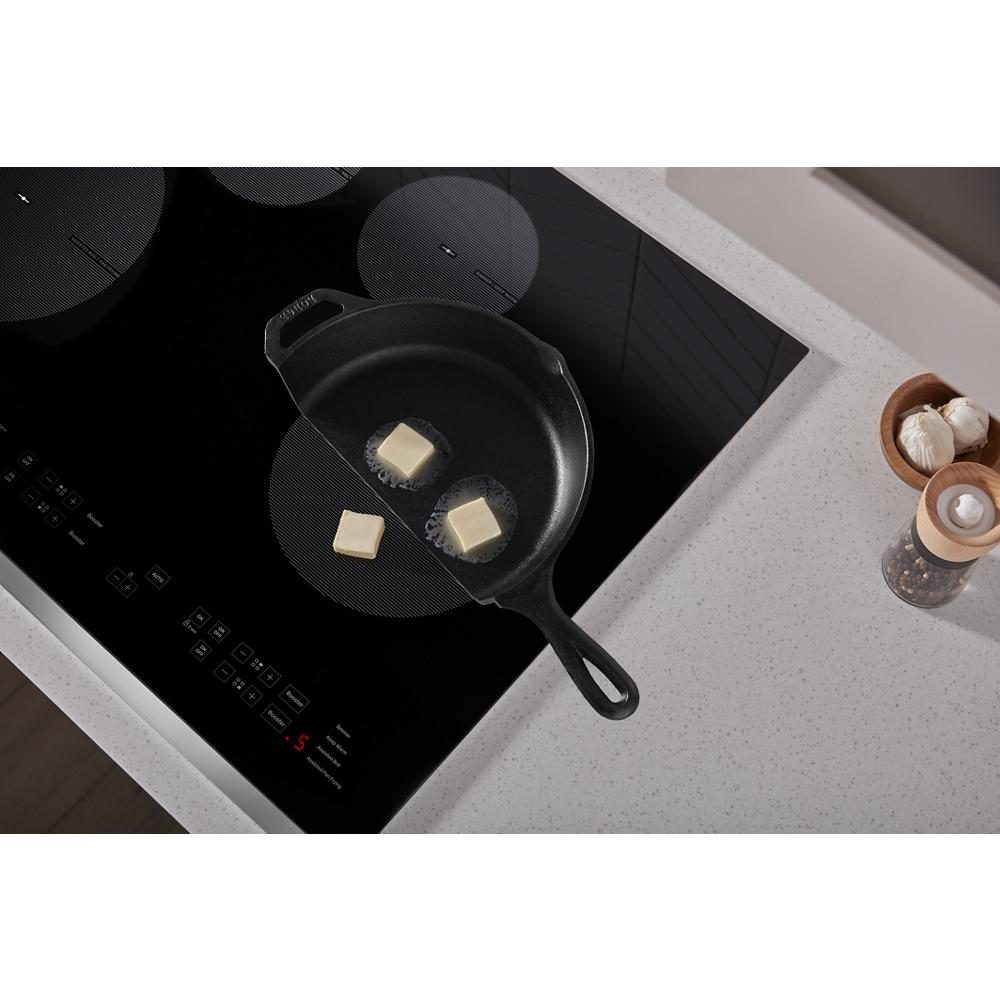 Black glass induction cooktop with cast iron pan melting butter on speckled white countertop in modern kitchen setting