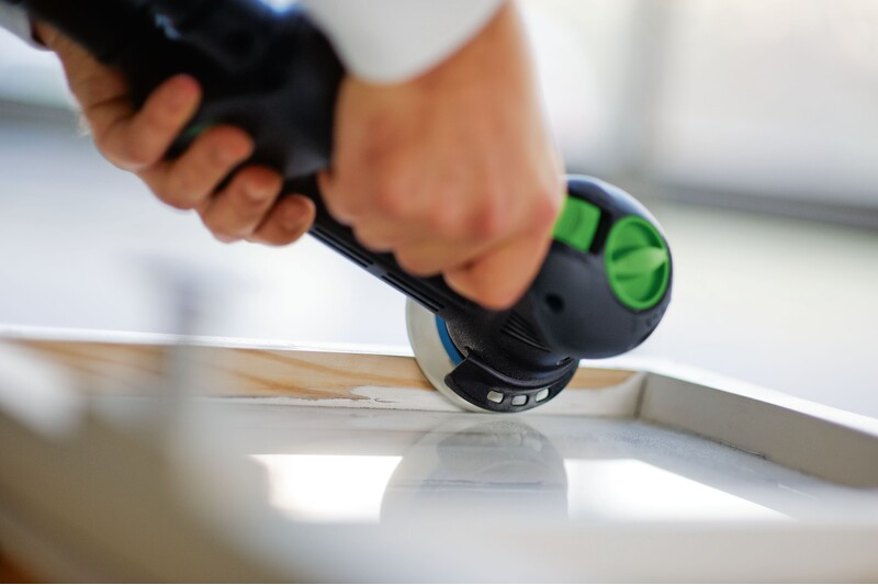 Person using a green and black multi-mode sander on a white wooden frame for home improvement tasks