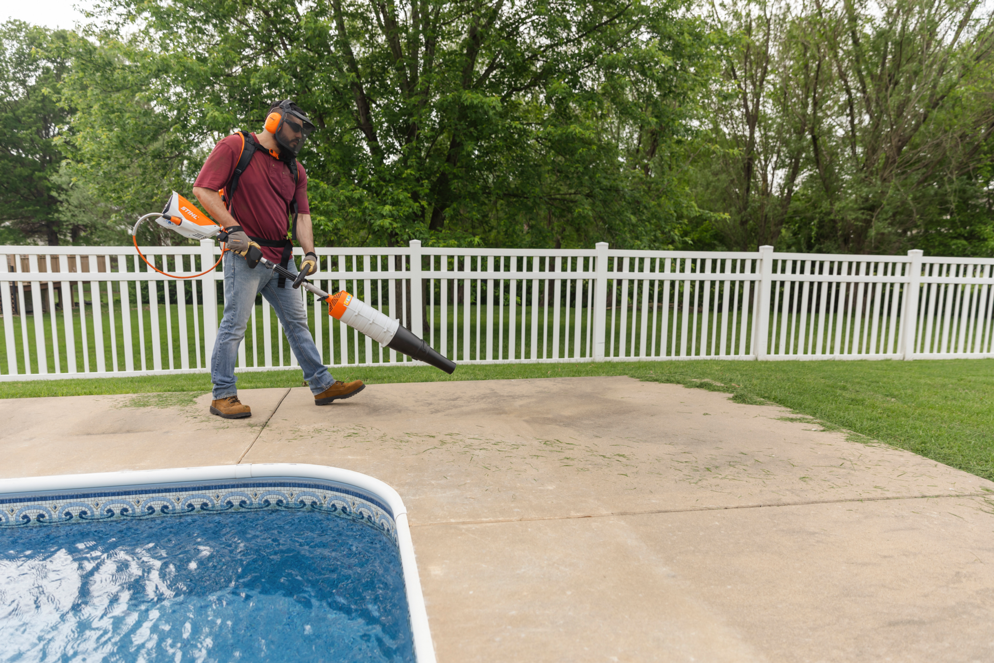 Man wearing protective gear using a battery-powered leaf blower on concrete near a pool in outdoor power equipment setting