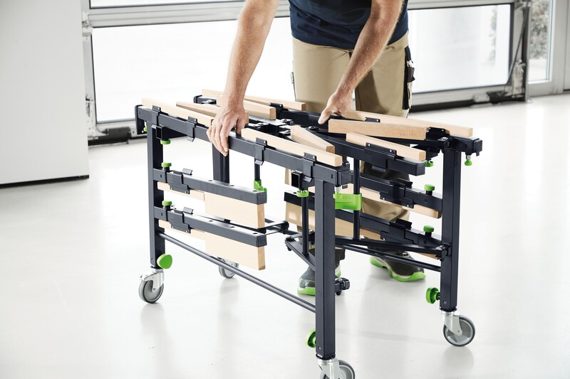 Person unfolding a black and wooden mobile workstation frame with locking caster wheels in a workshop setting