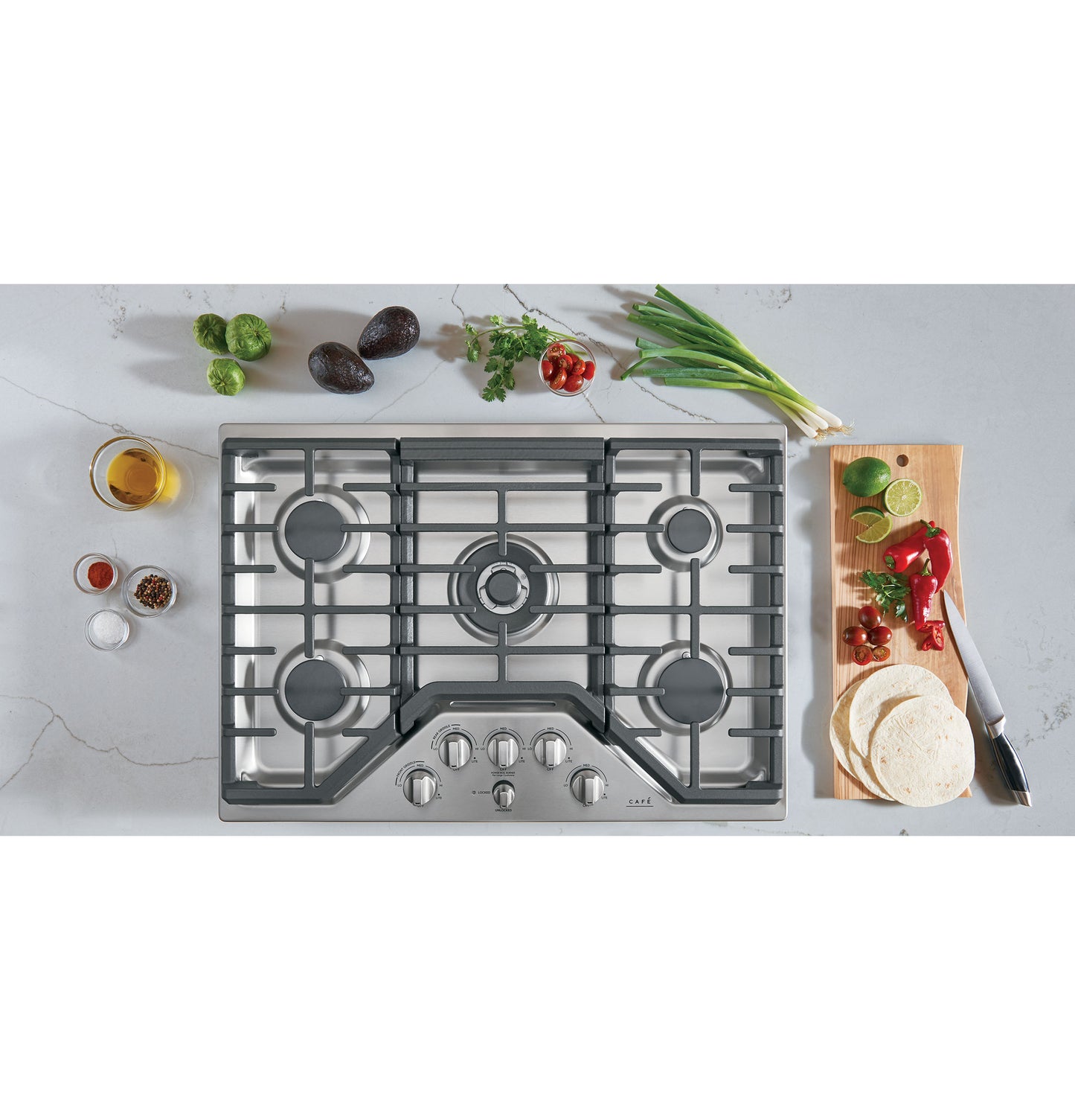 Stainless steel gas cooktop with five burners and front control knobs surrounded by fresh ingredients in kitchen setting