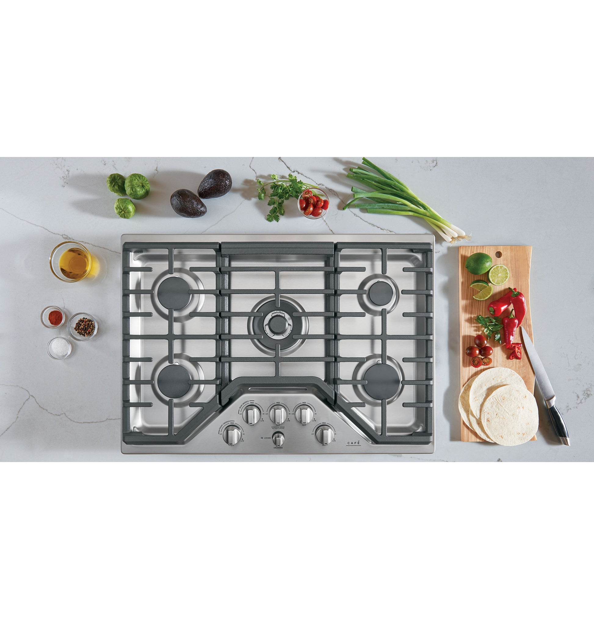 Stainless steel gas cooktop with five burners and front control knobs surrounded by fresh ingredients in kitchen setting