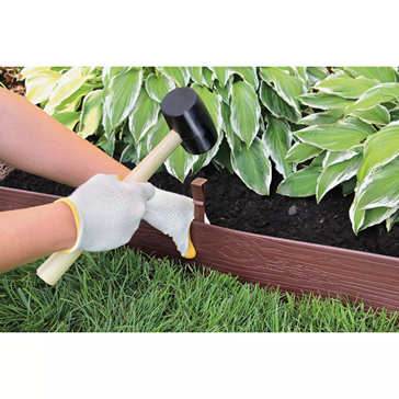 Person wearing gloves installing brown terrace board edging stakes with a rubber mallet along garden bed for home improvement tasks
