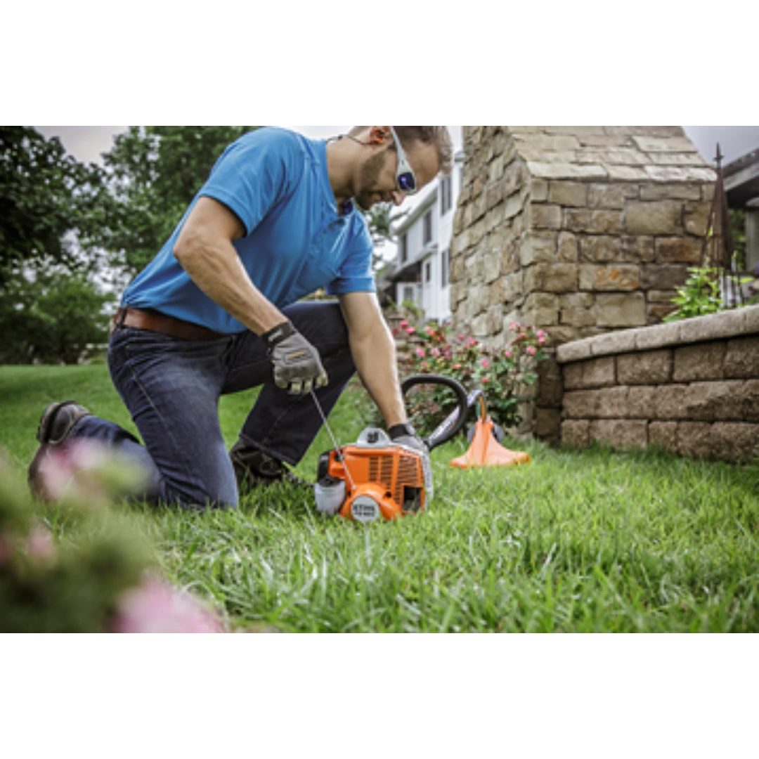 Man kneeling on grass adjusting orange gas-powered trimmer with curved shaft for yard maintenance tasks