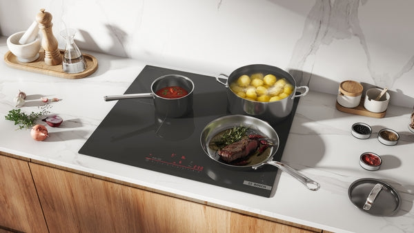 Black glass induction cooktop with three cooking zones heating pots of soup, potatoes, and steak in a modern kitchen setting