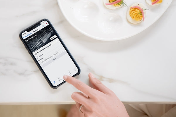 Hand interacting with smartphone displaying dishwasher status on app next to a plate of appetizers on marble countertop