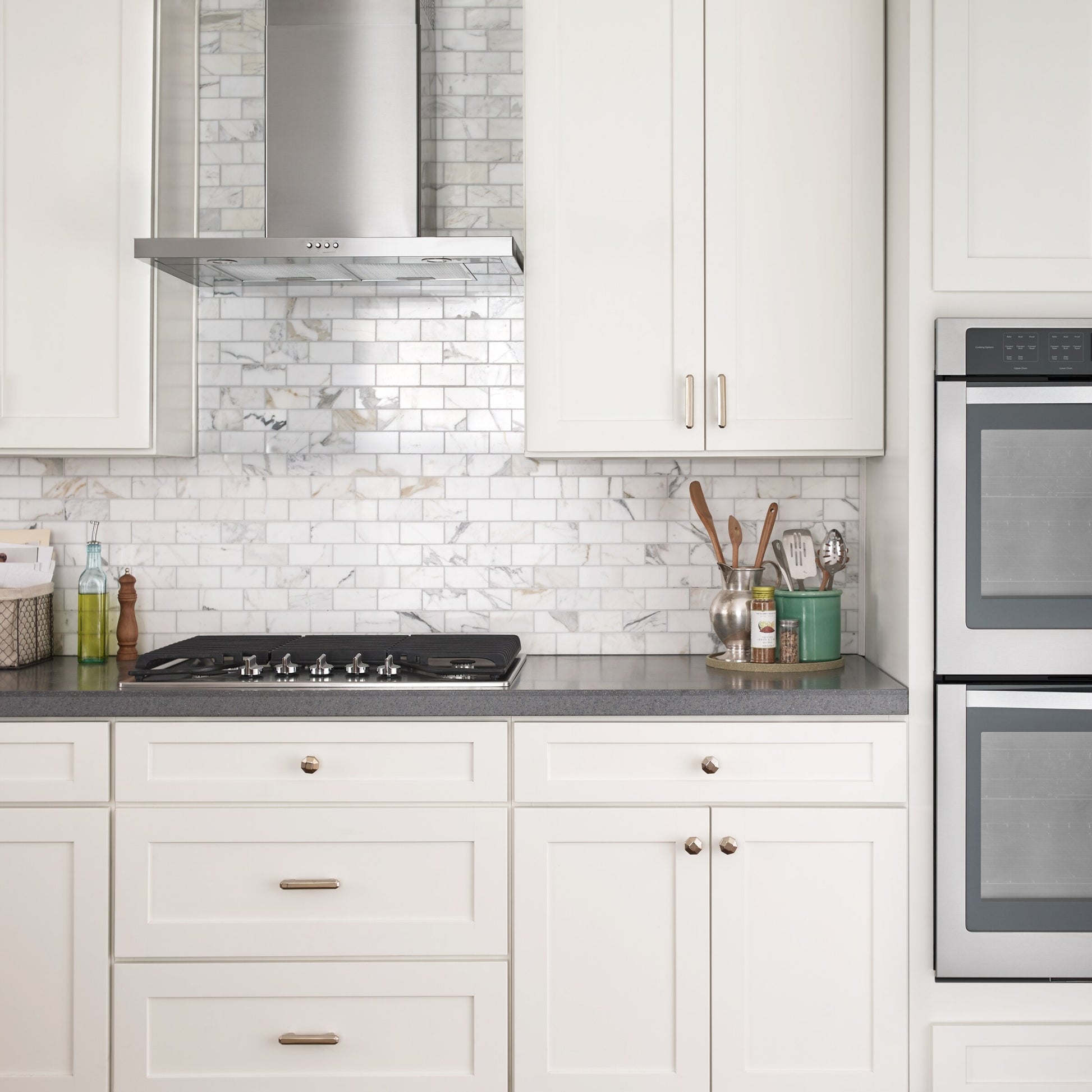 Stainless steel wall-mounted range hood above gas cooktop with white cabinetry and marble backsplash in modern kitchen
