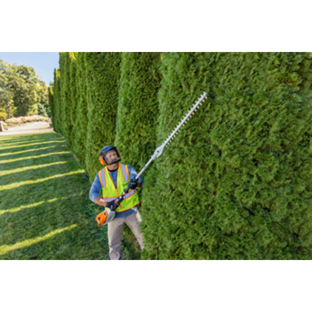 Worker wearing safety gear trimming tall hedge with extended-reach battery hedge trimmer for home improvement tasks