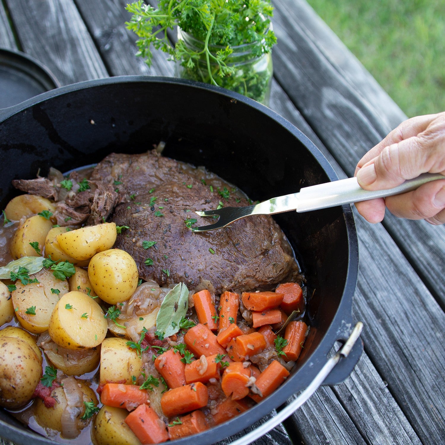 Hand holding stainless steel carving fork with white handle over pot roast with potatoes and carrots outdoors in kitchenware collection
