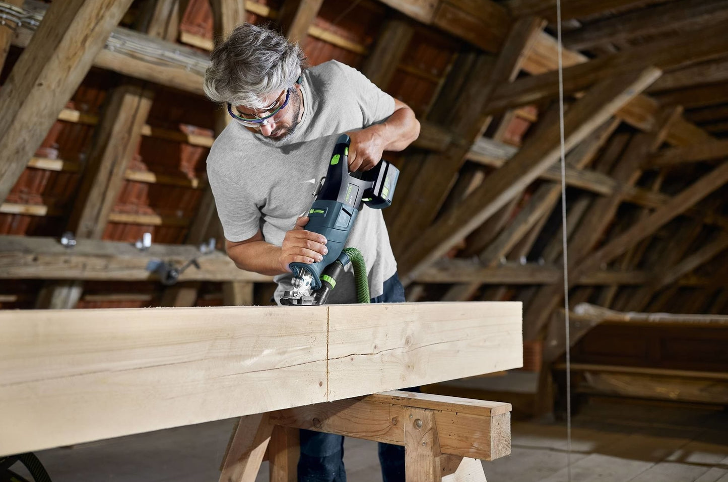 Man using Festool cordless reciprocating saw on large wood beam in a rustic attic home improvement setting