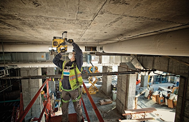 Construction worker wearing safety gear operating a cordless rotary hammer drill on a concrete ceiling at a job site