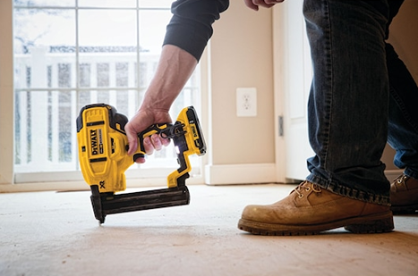 Person wearing brown boots using a yellow cordless narrow crown stapler on a wooden floor for home improvement tasks