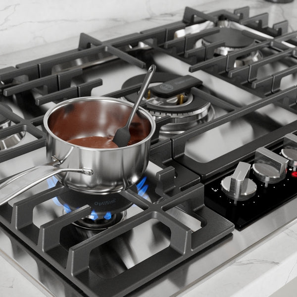 Stainless steel gas cooktop with blue flame heating a pot with chocolate batter, featuring cast iron grates and control knobs