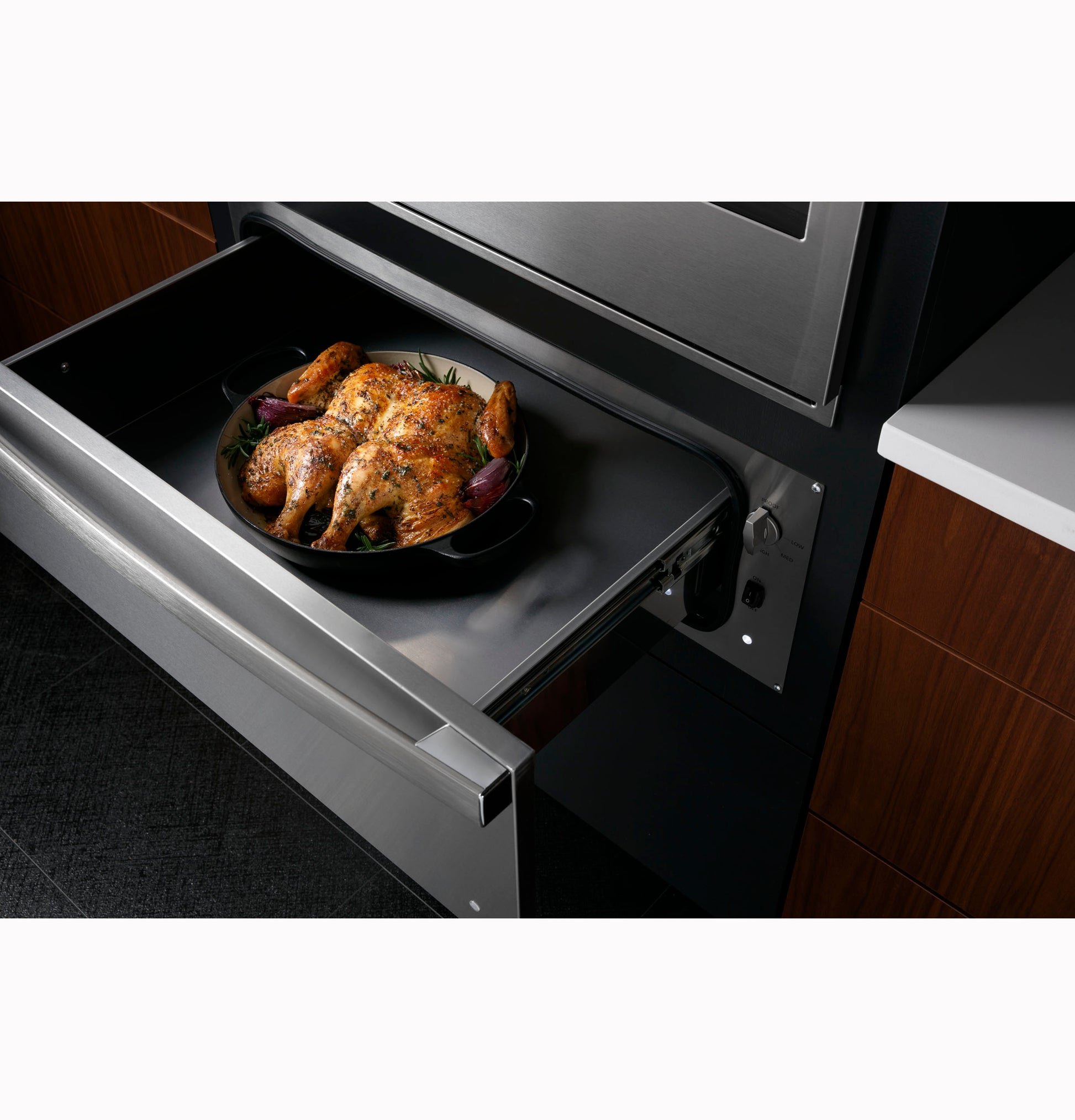 Open stainless steel warming drawer in modern kitchen with roasted whole chicken on a black dish inside appliance drawer