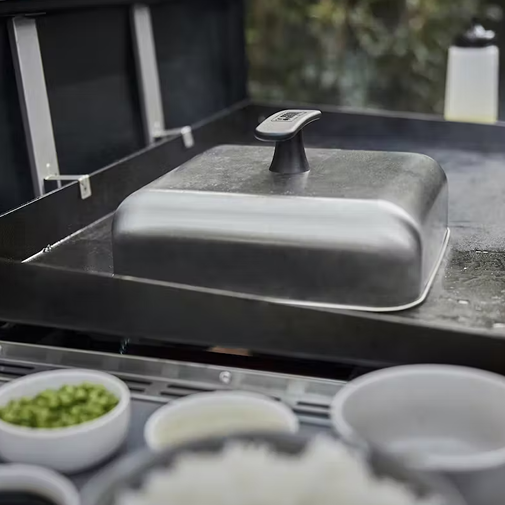 Stainless steel basting dome with black handle on a flat grill surface for outdoor cooking grilling accessories