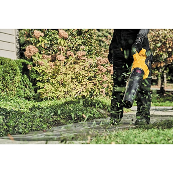 Person using a yellow cordless axial blower to clear grass clippings near a hedge in a garden setting