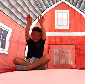 Child sitting inside a red inflatable barn play tent with gray roof and white window details raising arms