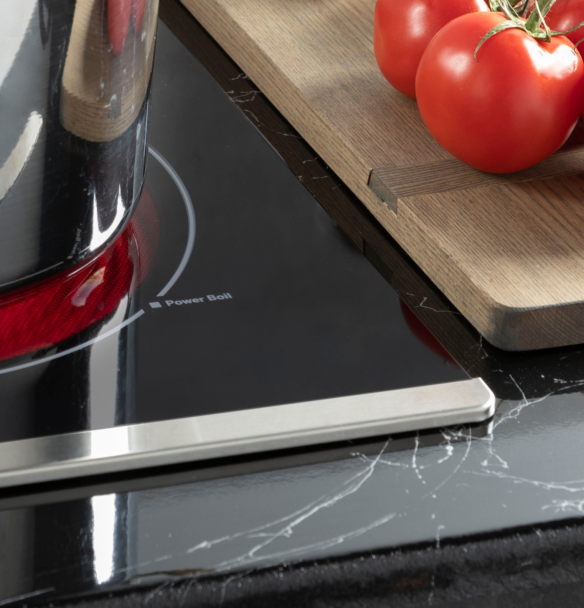 Close-up of black glass and stainless steel cooktop with active red heating element and fresh tomatoes on wooden cutting board
